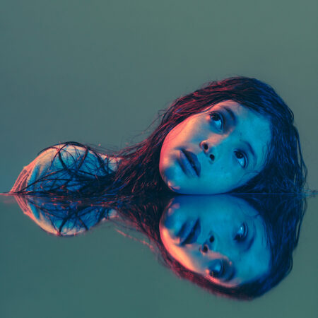 A young girl with long dark hair rests her head on a reflective surface and it looks like there are two of her. The background is green.